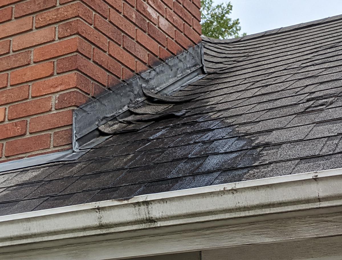 Damaged shingles and lifted flashing around a brick chimney